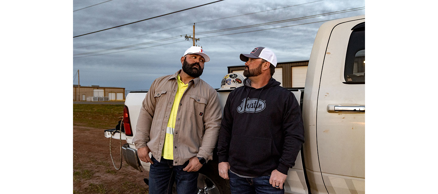 Two linemen, Kevin Walker and Dylan Edgmon, in workwear and ball caps stand beside a truck at dusk, talking casually near a job site with buildings and power lines in the background.